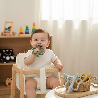Bébé souriant dans une chaise haute, mâchant un jouet de dentition vert en silicone de Petit Éveil Montessori.