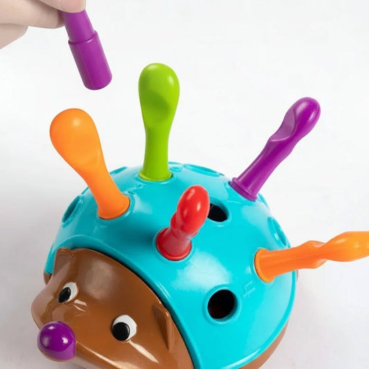 Petit Éveil Montessori : jouet hérisson bleu et marron avec des piquants colorés (orange, vert, rouge, violet) à insérer dans les trous pour développer la motricité fine.